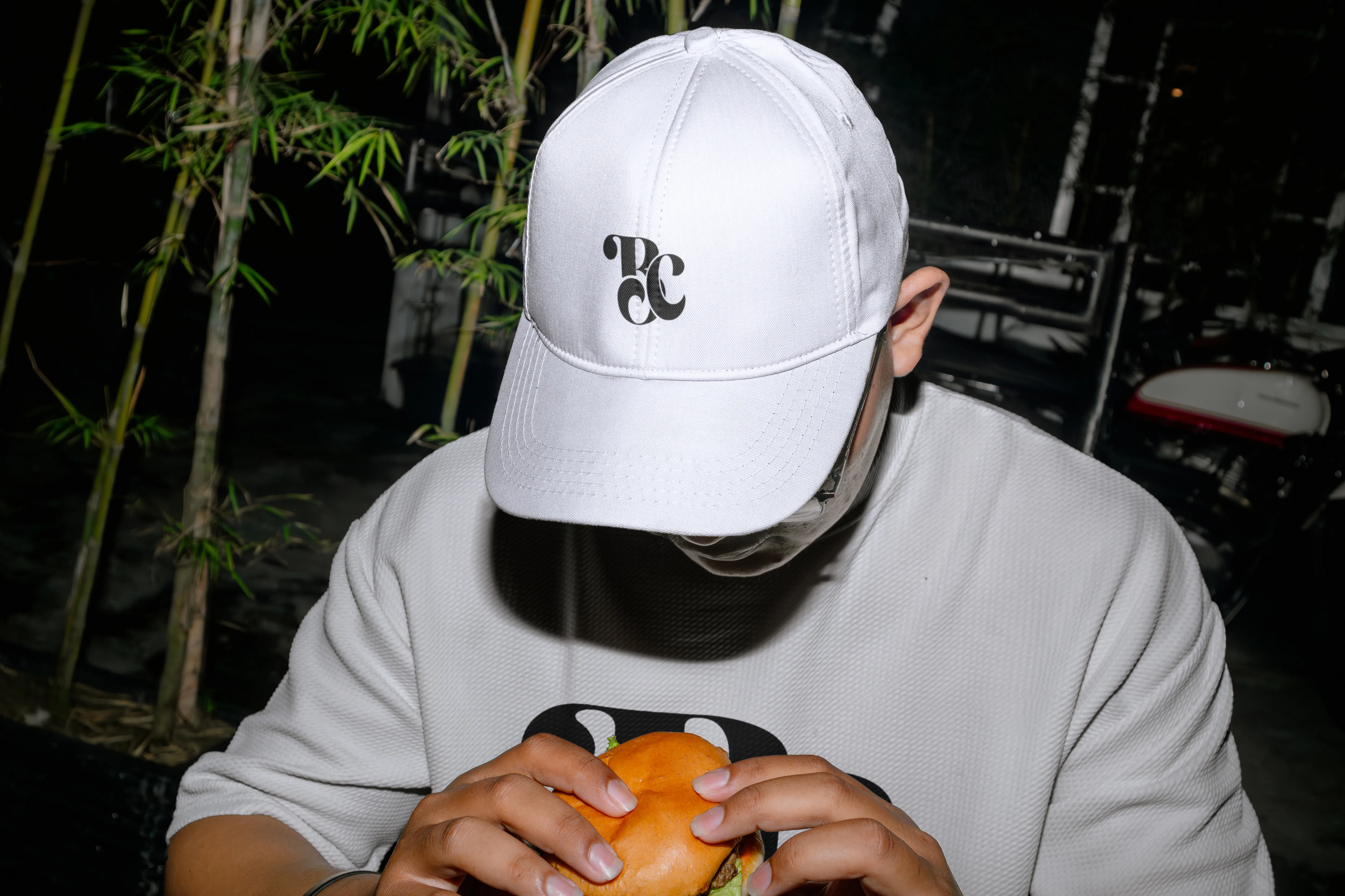Persona con gorra blanca personalizada mediante sublimación, con un logotipo estilizado en negro al centro, sosteniendo una hamburguesa.